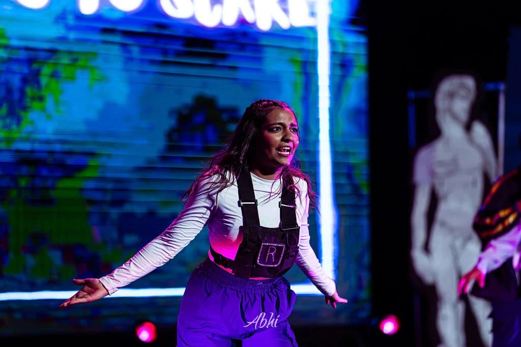 An NDDL performer hitting a sharp gesture beneath purple stage lights